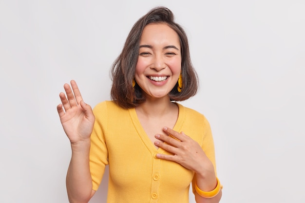 Feliz mujer asiática bonita de pelo oscuro con expresión positiva se ríe alegremente mantiene la mano levantada sonríe ampliamente usa aretes de puente amarillo oye algo divertido aislado sobre una pared blanca