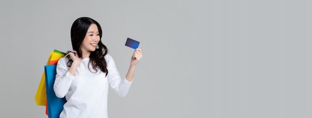 Feliz mujer asiática en camiseta blanca de manga larga mostrando tarjeta de crédito y llevando bolsas de compras aisladas