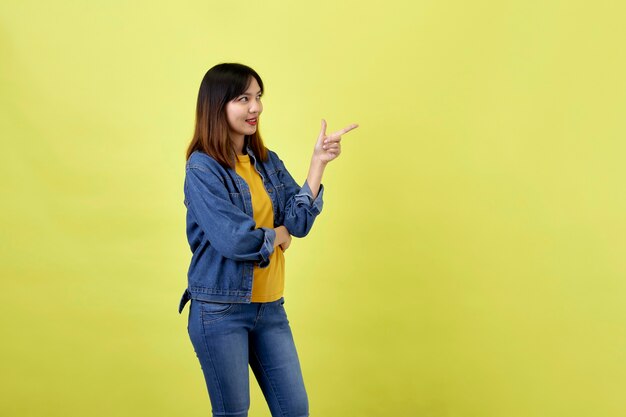 Feliz mujer asiática en chaqueta de mezclilla apuntando hacia el lado para copiar el espacio y mirando a la cámara sobre el espacio amarillo