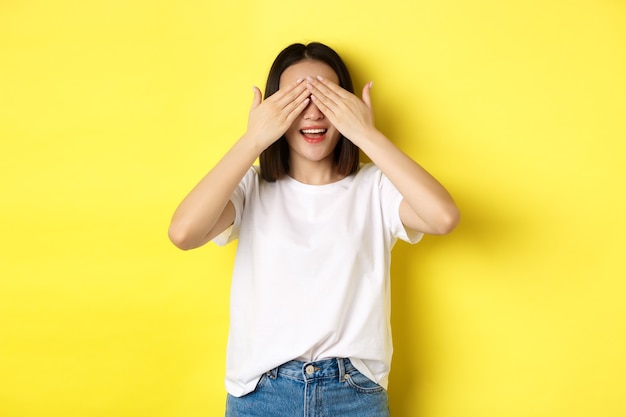 Feliz mujer asiática esperando un regalo sorpresa, cerrar los ojos y sonriendo, esperando algo, de pie sobre fondo amarillo