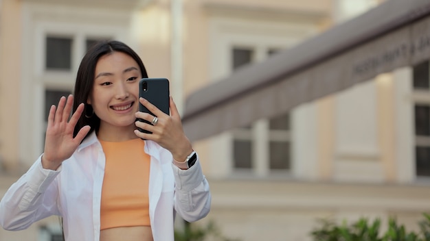 Feliz mujer asiática hablando por videollamada en la calle mujer sonriente haciendo videollamadas en el teléfono móvil en la ciudad a pie chica haciendo una videollamada sonriendo hablando agitando su mano