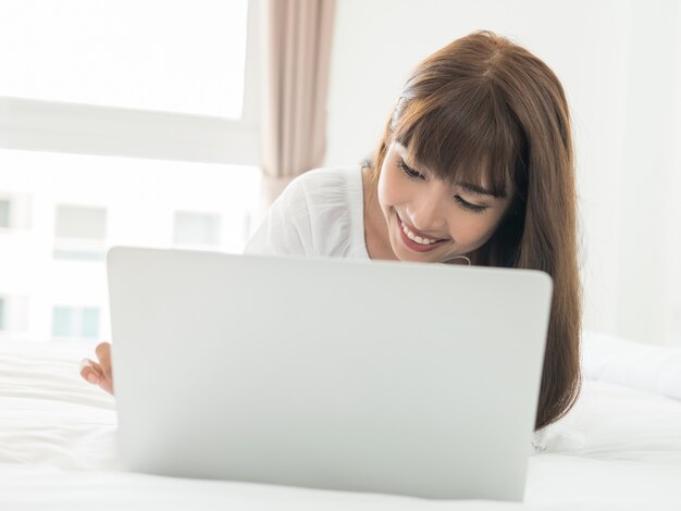 Feliz mujer asiática hermosa casual acostado en la cama usando la computadora portátil en el dormitorio en casa