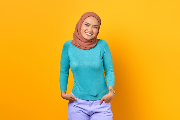 Feliz mujer asiática joven confiada sonriendo a una cámara con las manos en los bolsillos sobre fondo amarillo