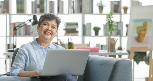 Feliz mujer asiática de mediana edad con pelo corto sonriendo y navegando por netbook mientras descansa en el sofá en la moderna sala de estar iluminada por el sol
