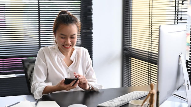 Feliz mujer asiática milenaria sentada en su lugar de trabajo y usando un teléfono móvil para comunicarse en las redes sociales