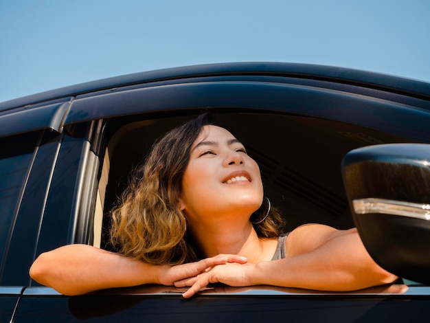 Feliz mujer asiática con pelo corto viajando en coche. Viajeros femeninos atractivos disfrutan y sonríen mientras miran la vista exterior con sol, verano.