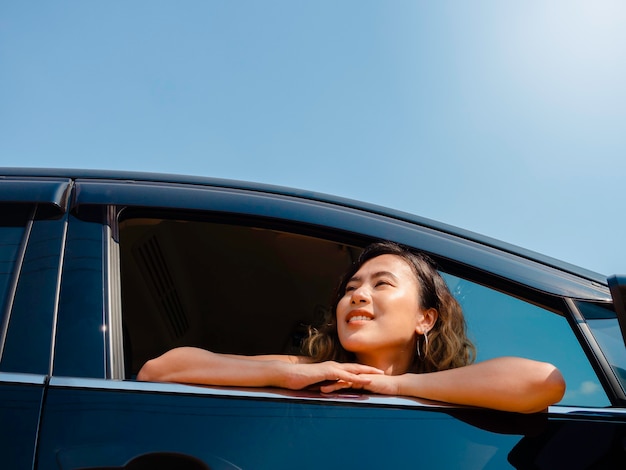 Feliz mujer asiática con pelo corto viajando en coche. Viajeros mujeres atractivas disfrutan y sonríen mientras miran la vista exterior sobre fondo de cielo azul con sol, verano.