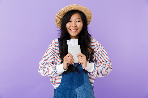 Feliz mujer asiática posando aislada sobre pared púrpura con pasaporte y boletos