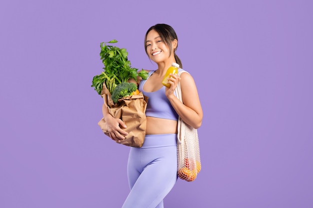 Feliz mujer asiática en ropa deportiva sosteniendo una bolsa con verduras y batido fresco