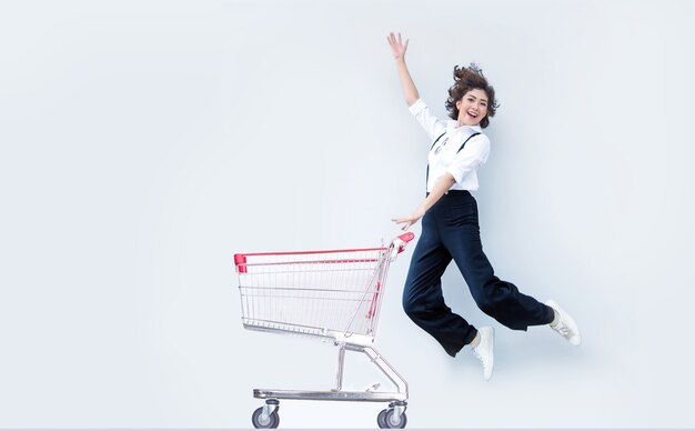 Feliz mujer asiática saltando con carrito de compras sobre fondo blanco.