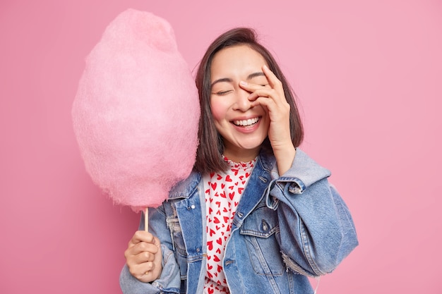 Feliz mujer asiática sincera tiene expresión despreocupada mano en la cara sonríe con alegría estar de buen humor sostiene hilo de caramelo dulce usa chaqueta de mezclilla elegante tiene tiempo libre expresa emociones positivas