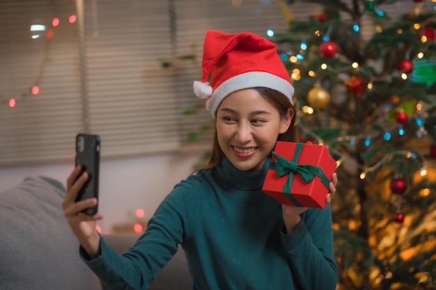 Feliz mujer asiática con smartphone tomando selfie móvil con caja de regalo de Navidad con árbol de Navidad en segundo plano.