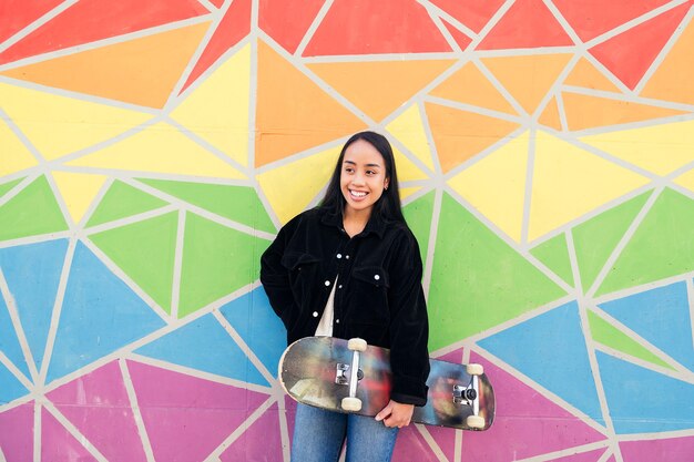 Feliz mujer asiática sonriendo apoyado contra una pared colorida con su patineta