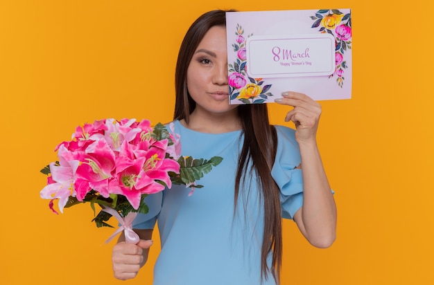 Feliz mujer asiática sosteniendo saludo cubriendo un ojo con tarjeta sosteniendo ramo de flores celebrando el día internacional de la mujer de marzo