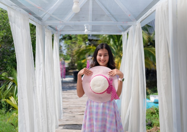 Feliz mujer asiática en vestido casual con sombrero en arco decorado con cortina blanca en jardín