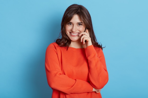 Feliz mujer de aspecto agradable con cabello oscuro hablando por teléfono, escuchando buenas noticias, se ve con una sonrisa encantadora, vistiendo un jersey naranja casual aislado sobre una pared azul, mujer hablando por teléfono celular