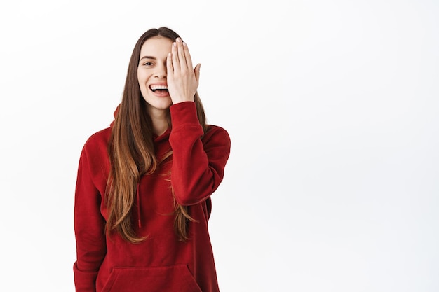 Feliz mujer atractiva con largo cabello castaño natural, cubrir la mitad de la cara con maquillaje desnudo, sonriendo complacida, mostrando el efecto antes y después, verificando la visión, mirando a la cámara, fondo blanco