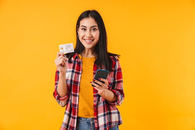 Feliz mujer atractiva sosteniendo una tarjeta de crédito y usando un teléfono celular aislado sobre una pared amarilla