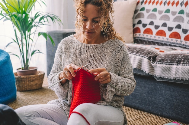 Feliz mujer bonita sonríe y admira su trabajo tejido con lana colorida roja. Las mujeres disfrutan de la actividad de ocio de tejido de interior en casa en la temporada de invierno