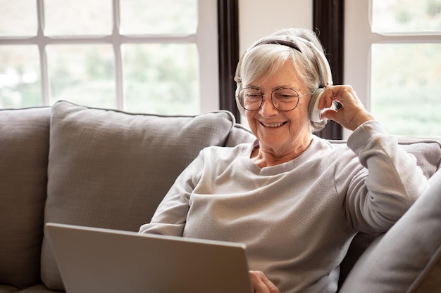 Feliz mujer de cabello blanco de los años 70 sentada en el sofá en la sala de estar sostiene una computadora portátil viendo una película Concepto de usuarios de tecnología inalámbrica y generación anterior