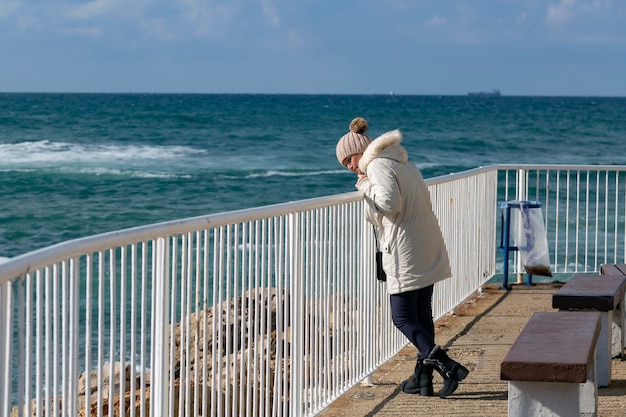 Feliz mujer caucásica americana de pie en la playa con una chaqueta blanca y gorro de punto con vistas al mar azul y disfrutando de la libertad. Mujer emocional caminando sobre el muelle blanco contra el mar