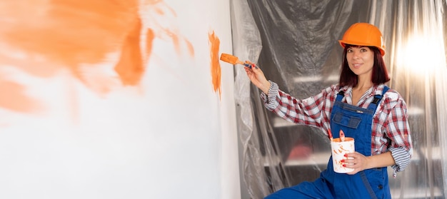 Feliz mujer caucásica con casco pinta la pared interior de una casa nueva con un pincel en naranja Concepto de construcción y renovación Renovación del hogar Fondo y lugar para texto Banner