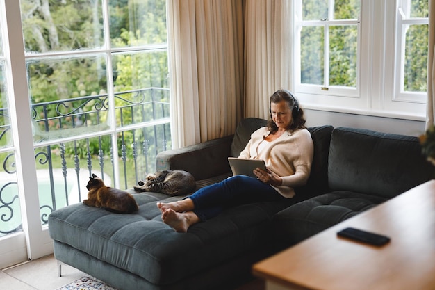 Feliz mujer caucásica mayor en la sala de estar sentada en el sofá, usando audífonos y usando una tableta