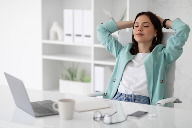 Feliz mujer caucásica del milenio en traje con los ojos cerrados descansando en el lugar de trabajo con una taza de té y