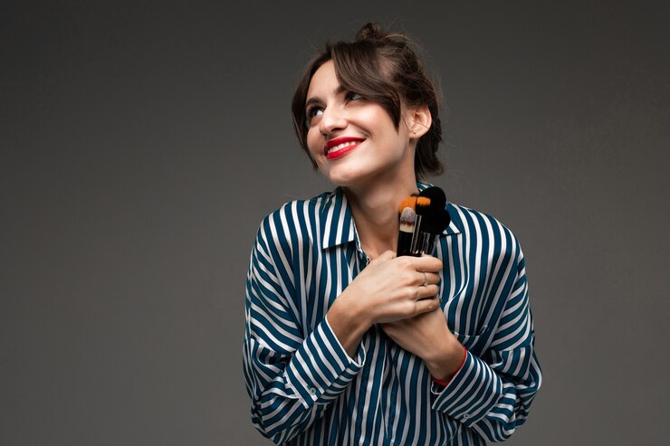 Feliz mujer caucásica tiene pinceles de maquillaje aislados en la pared gris
