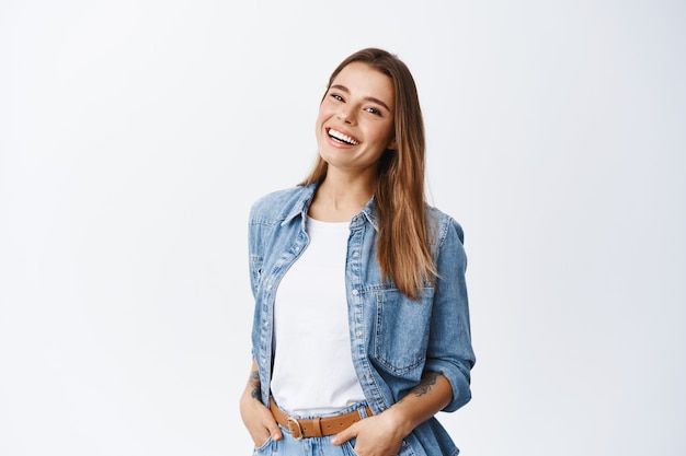 Foto feliz mujer despreocupada con expresión alegre, sonriendo mientras está de pie en pose relajada contra la pared blanca, tomados de la mano en los bolsillos de los pantalones vaqueros