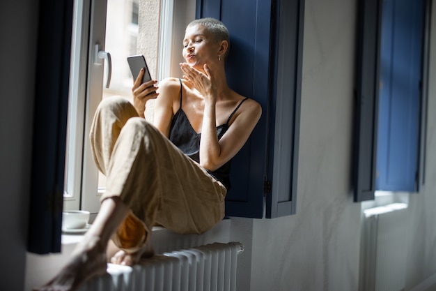 Feliz mujer elegante diviértase comunicándose por teléfono o haciendo una foto selfie mientras está sentada relajada en el alféizar de la ventana en casa.