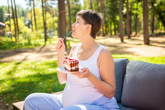 Feliz mujer embarazada comiendo pastel dulce en el parque