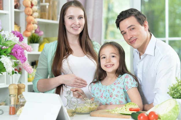 Feliz mujer embarazada con esposo e hija preparando ensalada juntos en la cocina
