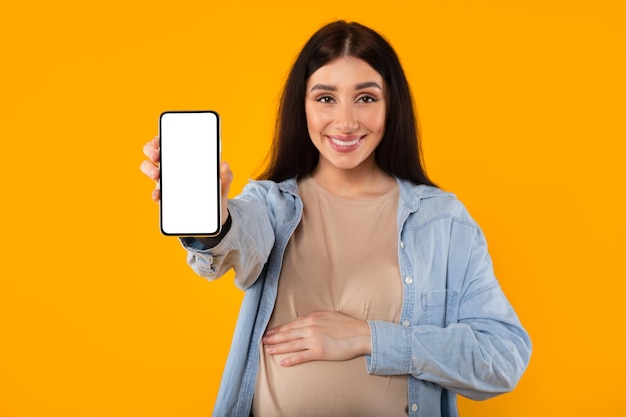 Feliz mujer embarazada europea con gran barriga mostrando teléfono inteligente con pantalla blanca en blanco en amarillo