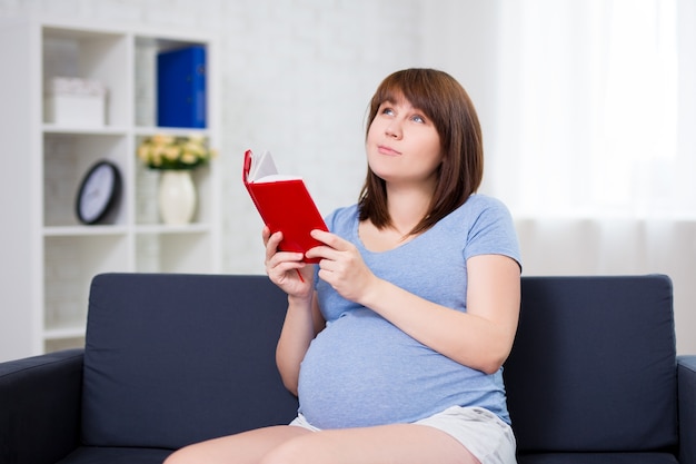 Feliz mujer embarazada leyendo el libro y pensando o soñando con algo en casa