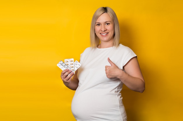 Feliz mujer embarazada, rubia hace un gesto de pulgar hacia arriba mientras sostiene las píldoras. medicamentos seguros para el embarazo