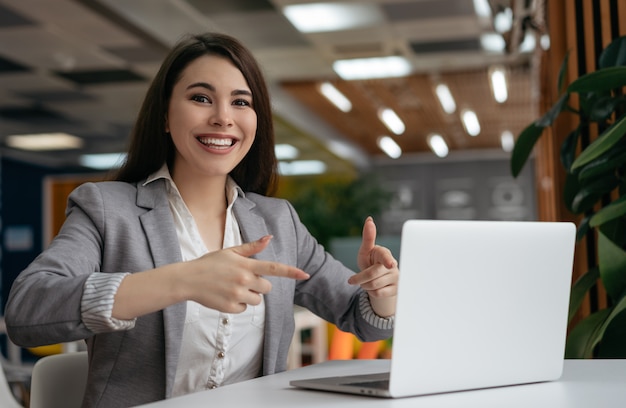 Feliz mujer emocional mostrando los dedos en la computadora portátil, mirando a cámara y sonriendo