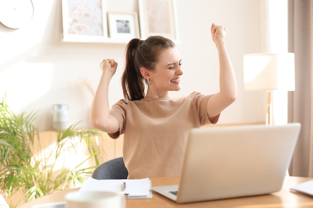 Feliz mujer emprendedora sentarse en el escritorio leyendo buenas noticias y expresar alegría levantando las manos.