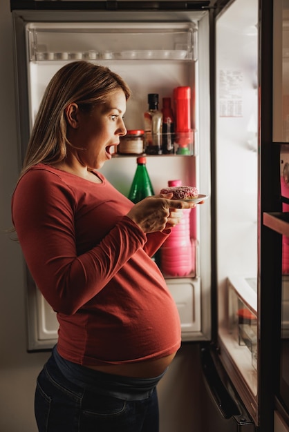 Feliz mujer esperando comer donas frente al refrigerador abierto en la cocina por la noche.