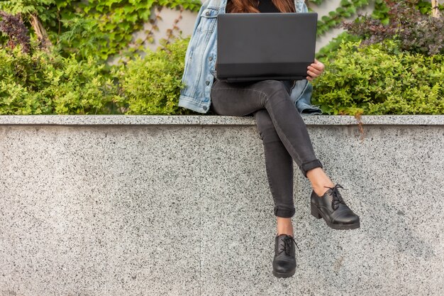 Feliz mujer estudiante en chaqueta vaquera y gafas usa laptop mientras está sentado en el parque de la ciudad