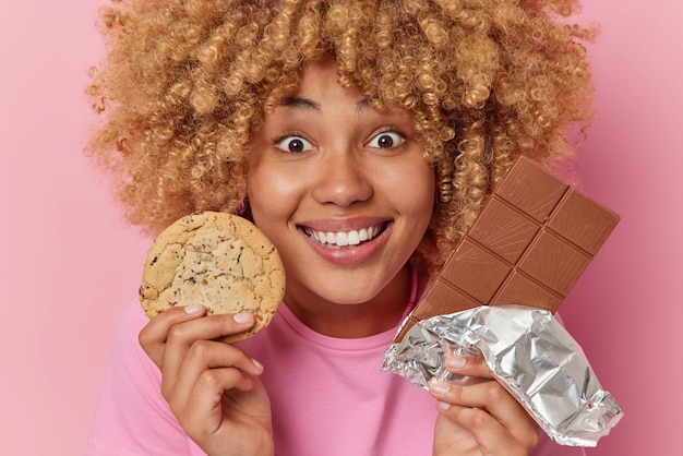 Feliz mujer europea de pelo rizado sostiene una barra de chocolate y una galleta disfruta de la dulce sonrisa de la vida felizmente prefiere comer comida azucarada poco saludable vestida con una camiseta informal aislada sobre un fondo rosa