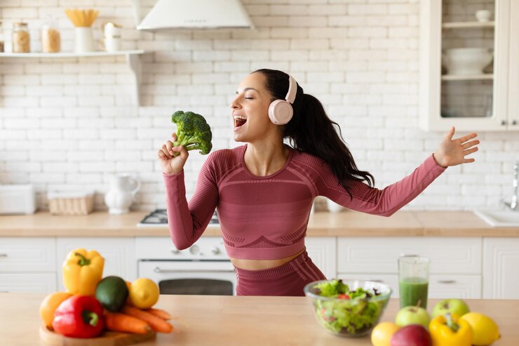 Foto feliz mujer fitness con auriculares cantando sosteniendo brócoli en la cocina