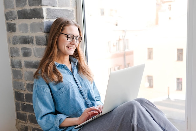 Feliz mujer hermosa casual con gafas trabajando en una laptop sentada en el alféizar de la ventana