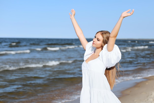 Feliz mujer hermosa sonriente en la playa del océano de pie en un vestido blanco de verano, levantando las manos.