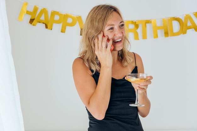 Foto feliz mujer hermosa en vestido negro bebiendo champán hablando por teléfono