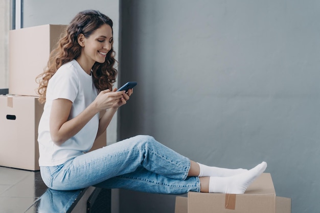 Feliz mujer hispana atractiva está sentada en el alféizar con cajas y mensajes de texto en el teléfono móvil