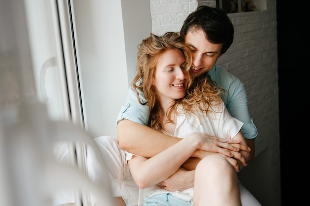 Feliz mujer y hombre abrazando junto a la ventana en casa el día de San Valentín