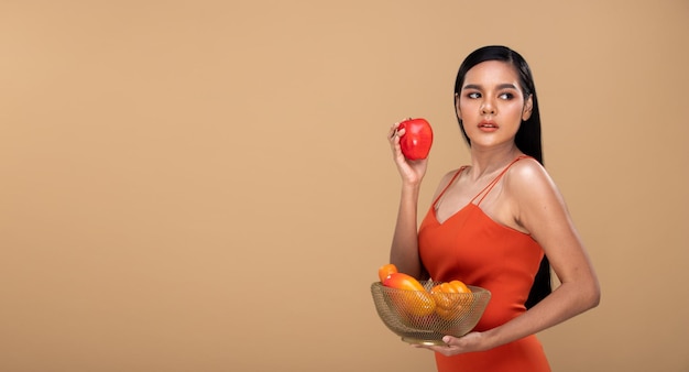 Feliz mujer india asiática con el pelo largo y negro y una gran sonrisa de dientes sosteniendo Apple en la mano y una cesta con frutas frescas saludables y una sonrisa de dientes blancos. Espacio de copia de fondo amarillo beige