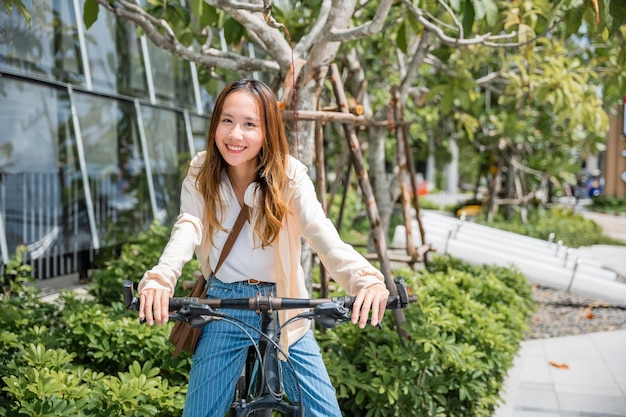 Foto feliz mujer joven asiática montando en bicicleta en la calle al aire libre cerca de la construcción de la ciudad, retrato de un estilo de vida femenino sonriente usando bicicleta en medios de transporte de viajes de verano, respetuoso con el medio ambiente