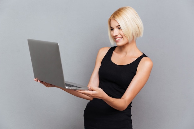 Feliz mujer joven y bonita sonriendo y usando la computadora portátil sobre pared gris